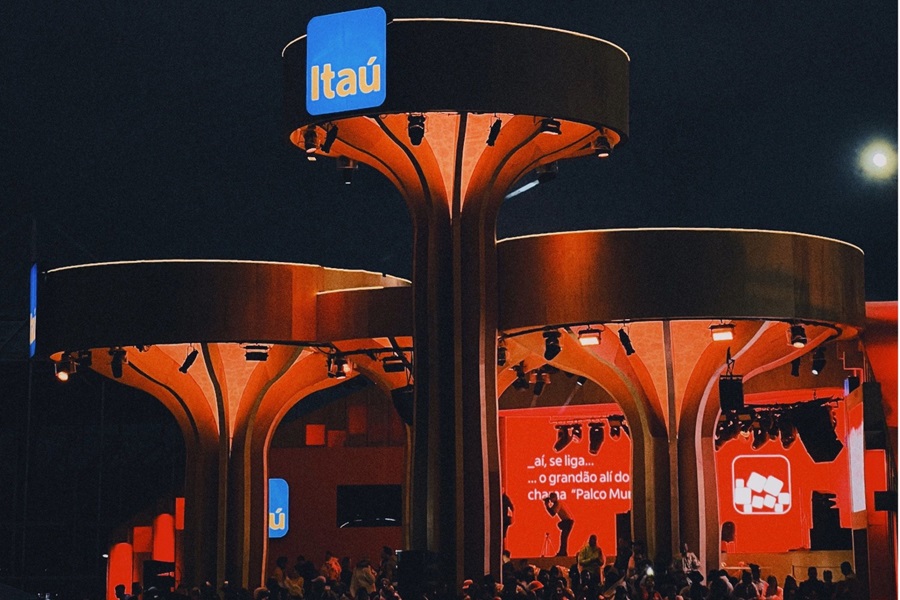 Illuminated Itaú-branded event space with striking orange-lit canopy structures, a stage with red screens, and an audience gathered below.