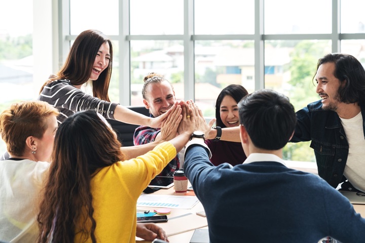 A group of seven people in a meeting, all doing a high five over the centre of the table together.