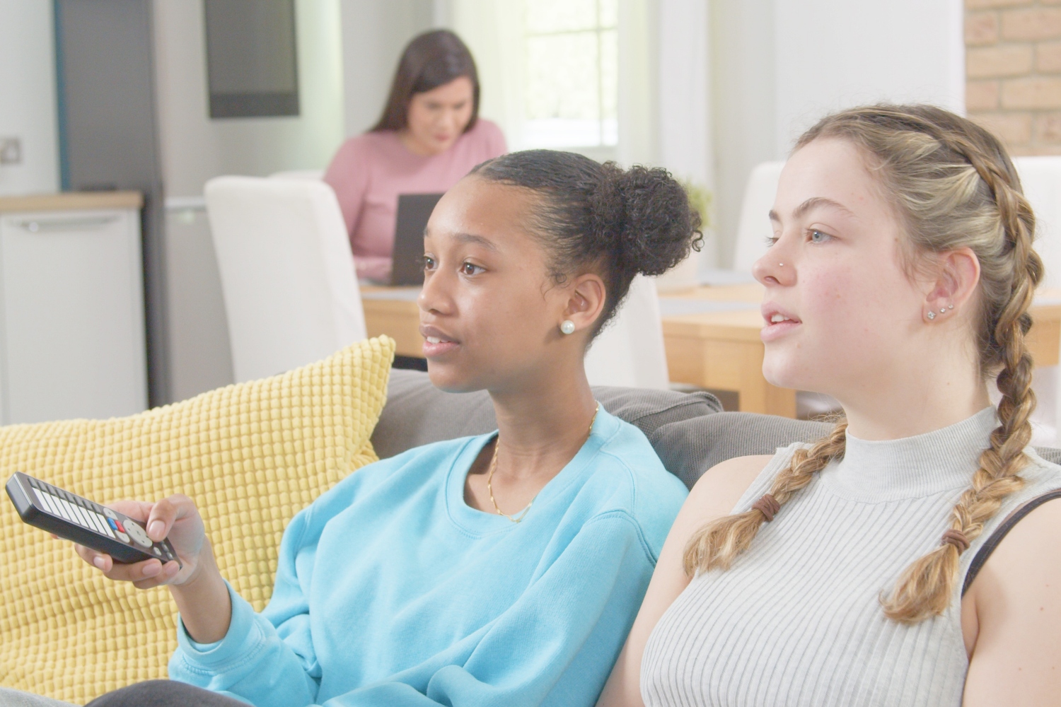 Two girls sat watching TV with a parent working on a laptop in the background