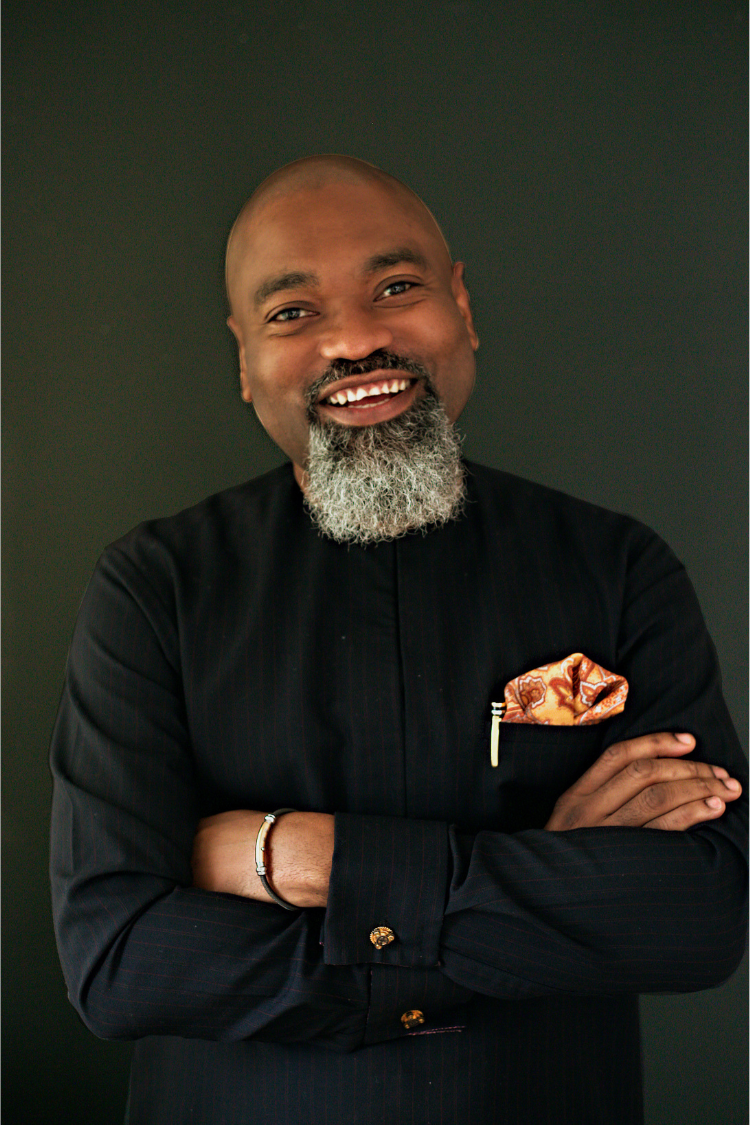 A torso upwards photograph of Adeola in a black shirt and orange pocket square with his arms folded. 