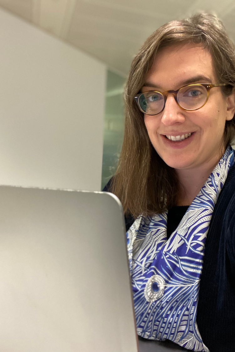 A woman working at a laptop, wearing a white and blue patterned short. She has mid length brown hair and is wearing glasses.