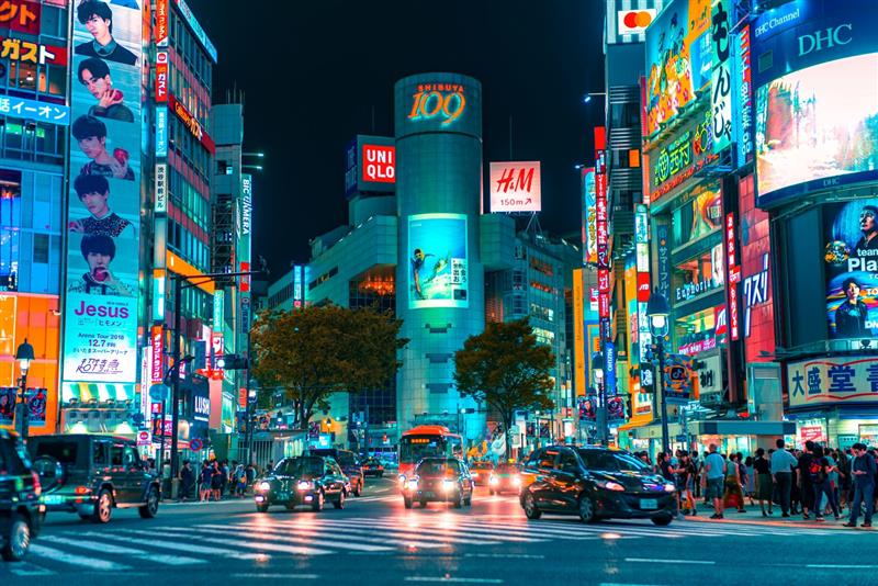 Neonlit Shibuya crossing at night with bright billboards, busy traffic, and crowds in Tokyo.