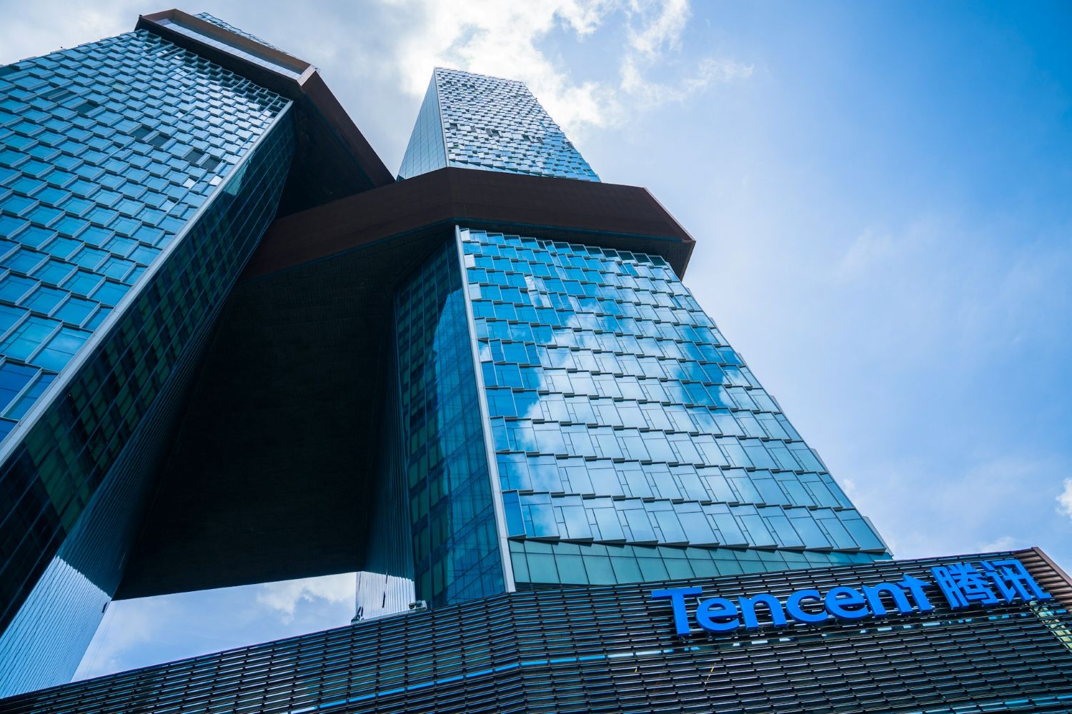 Tencent headquarters, a modern blue-glass skyscraper with interconnected towers, viewed from below against a bright sky.