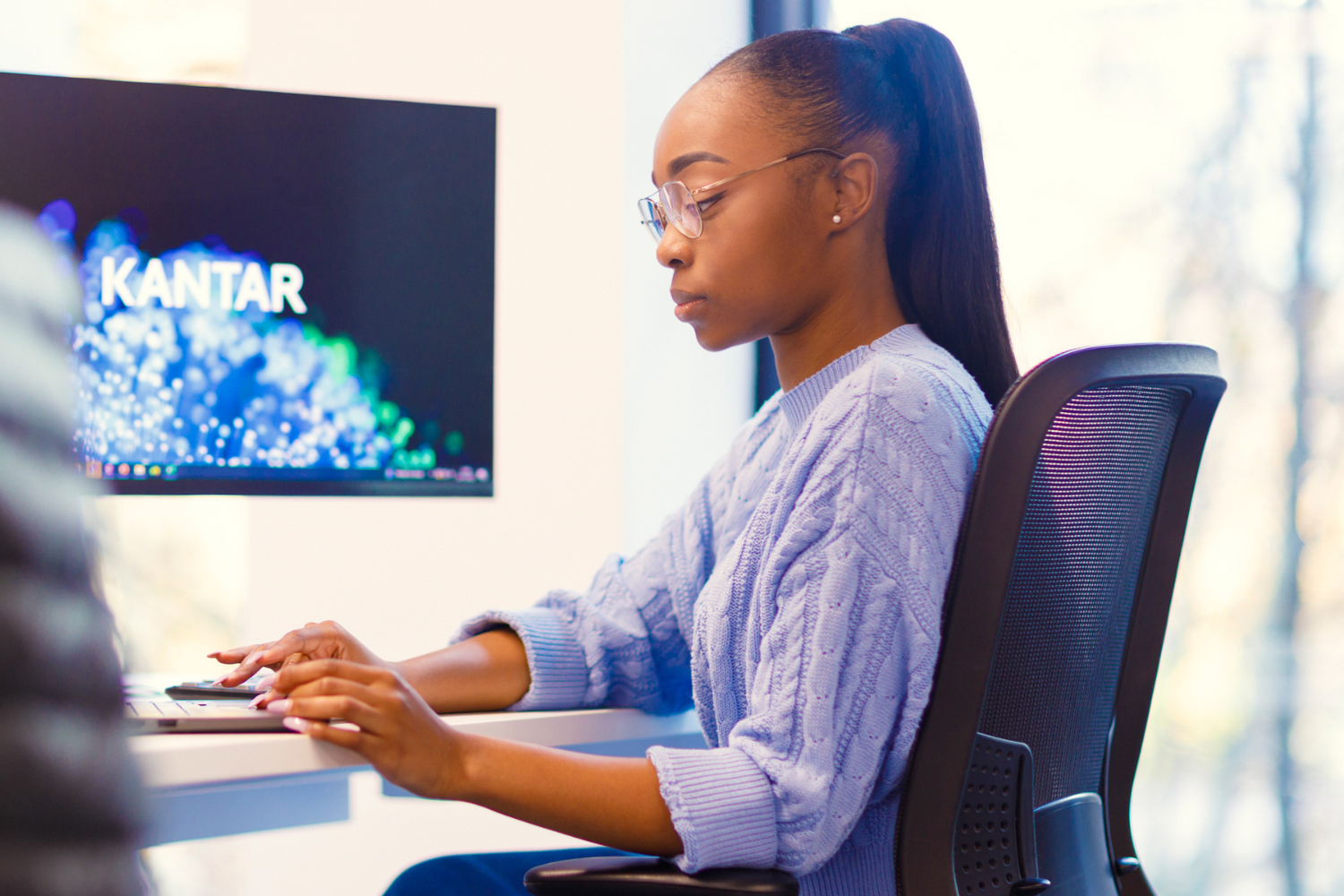 A tound girl is sat working her desk. She is wearing a pale blue top and has her hair in a high pony tail. 