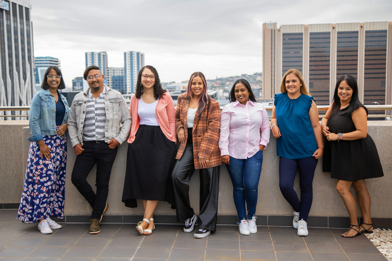 Seven casually dressed individuals standing on a rooftop with a city skyline in the background. From left to right: one wears a blue floral dress and denim jacket; another wears striped shirt and denim jacket; the third wears an orange cardigan over a white top; the fourth wears an orange plaid jacket; the fifth wears a pink shirt and jeans; the sixth wears a teal sleeveless top and dark jeans; and the seventh wears a short black dress. All are leaning against a railing under an overcast sky.