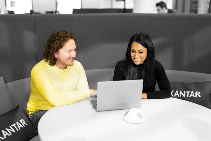 A man and a woman sat at a table with a laptop, working together. She has long dark hair and  a black top, he has brown curly hair and a  yellow jumper