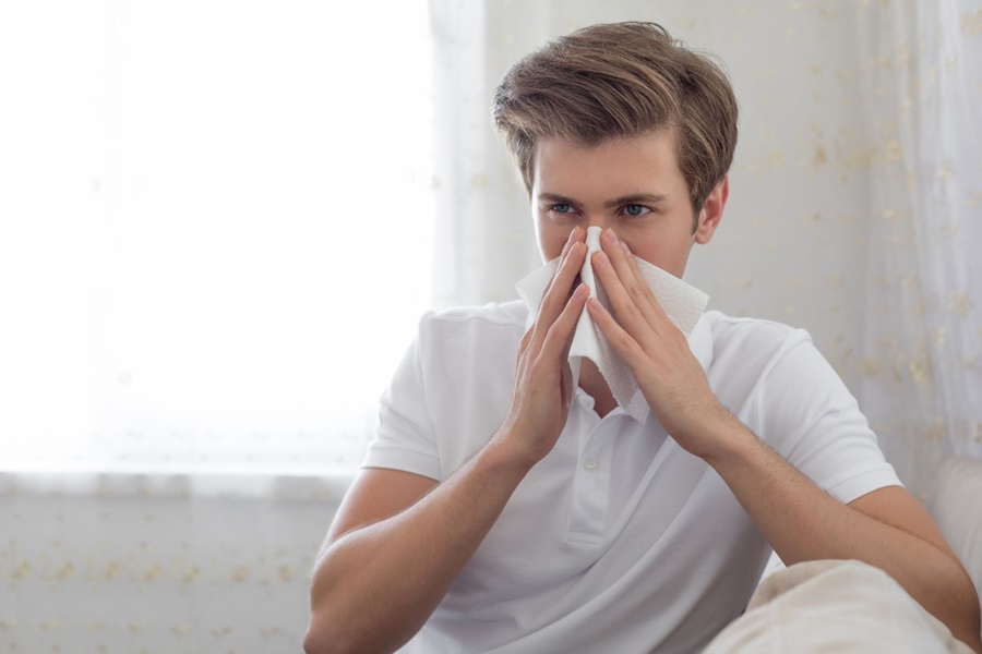 Picture of a man sat on sofa at home blowing nose on a tissue