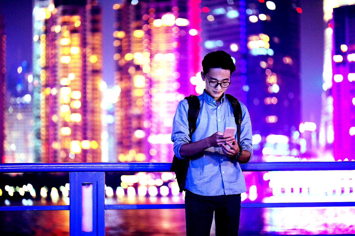 Man using smartphone near a river and skyscrapers in background.